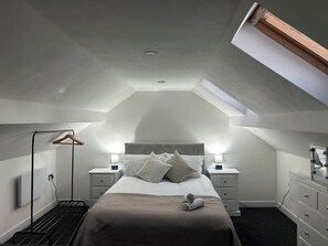 A bedroom with a sloped ceiling, a bed with pillows, bedside tables, a hanging clothes rack, and a dresser with a mirror.