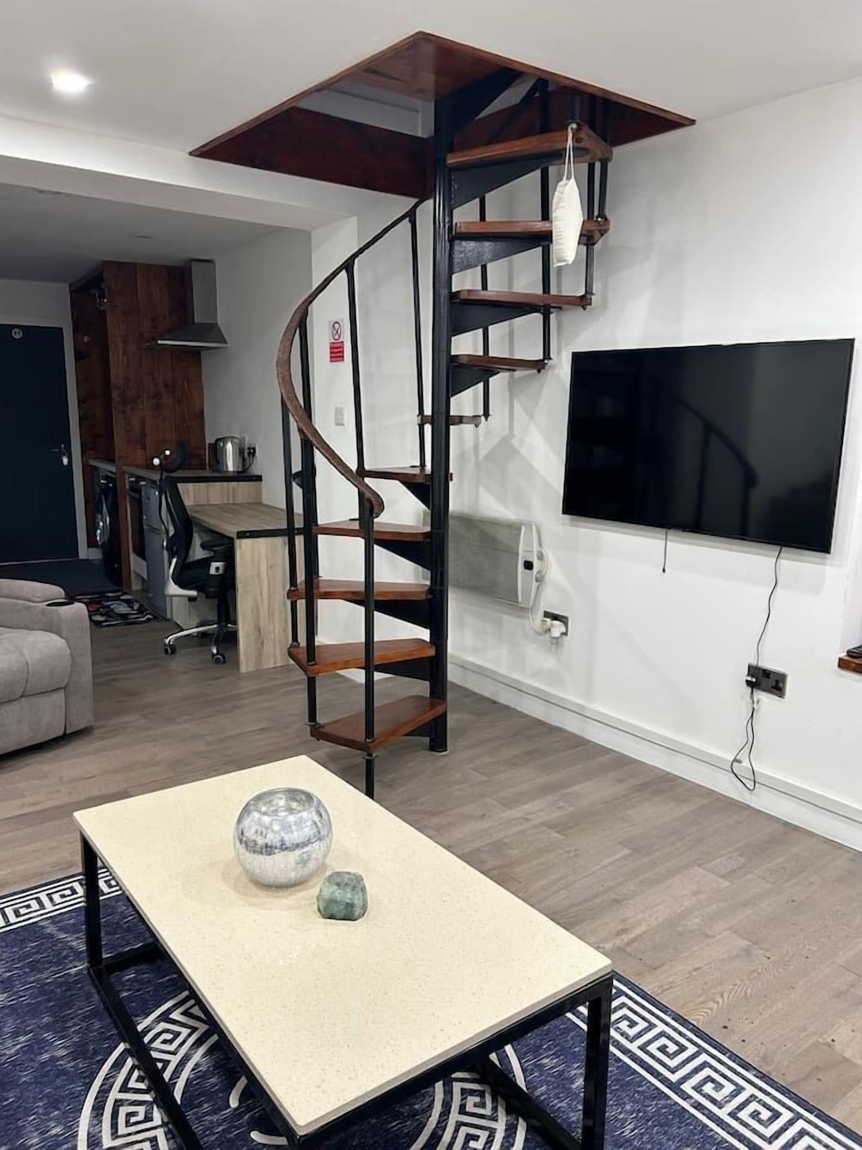 A modern living room with a spiral staircase, a flat-screen TV mounted on the wall, and a small table with decorative items.