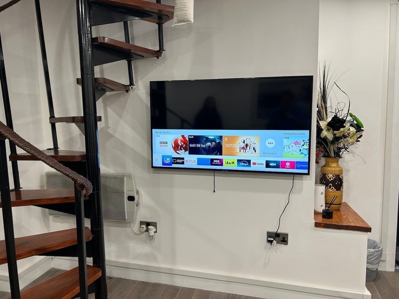 Televisor de pantalla plana montado en una pared blanca, con una escalera de madera a la izquierda.