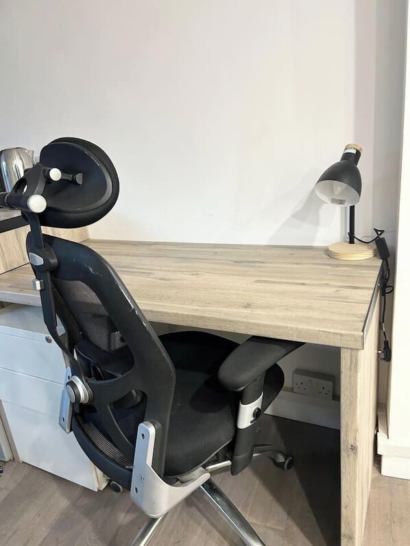 A wooden desk with a black office chair, a lamp, and a power outlet.