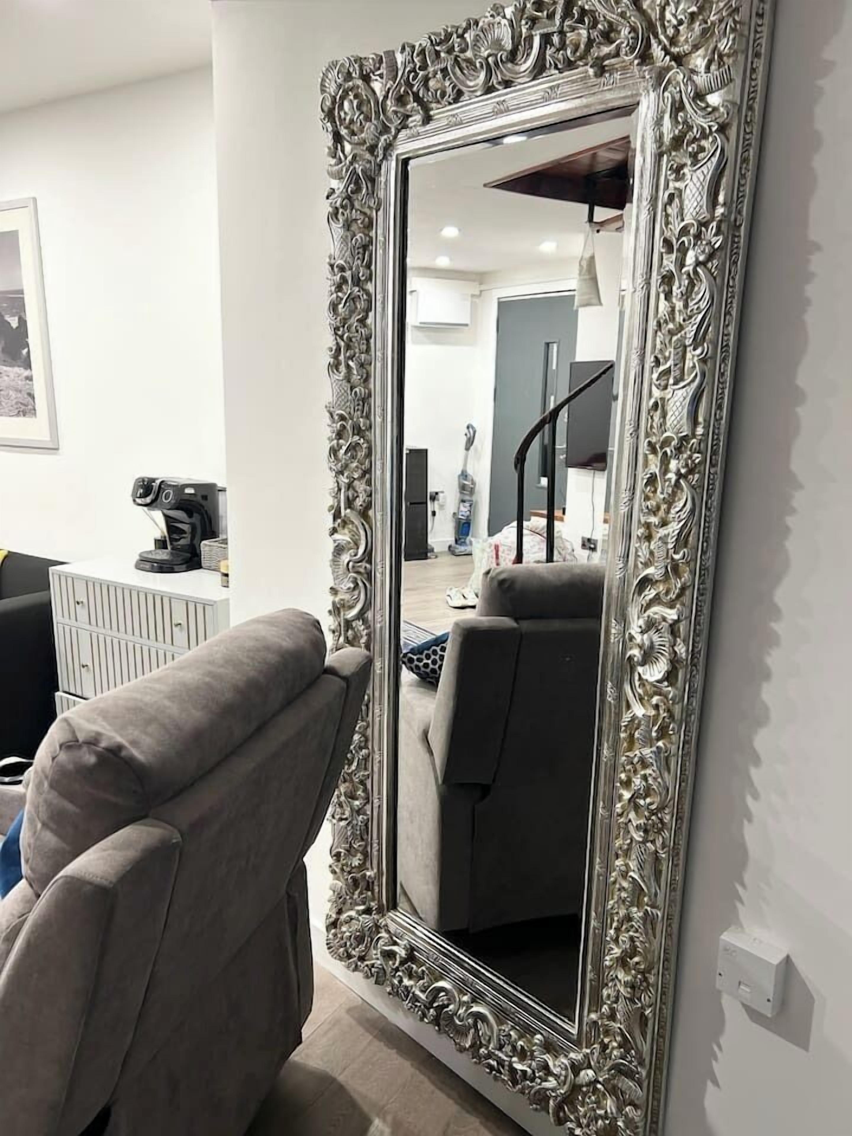 A room with a large ornate mirror, a grey armchair, and a white wall.