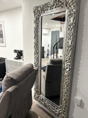 A room with a large ornate mirror, a grey armchair, and a white wall.