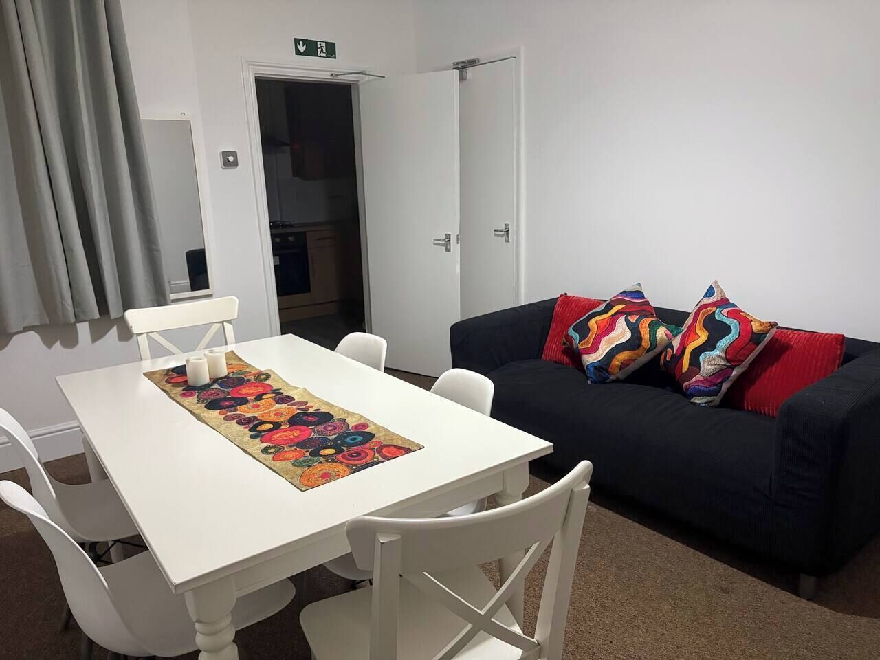 A room with a white table, white chairs, a black sofa with colorful pillows, and a door leading to another room.
