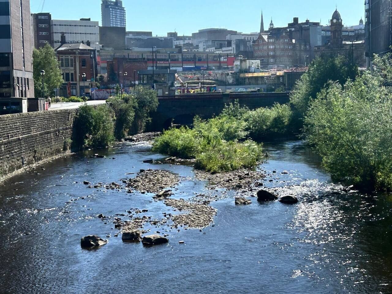 Un río con rocas, vegetación, un puente y edificios al fondo.