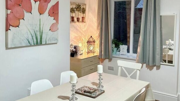 En matplats med ett bord dukat för fyra personer, en tavla på väggen och ett fönster med gardiner.