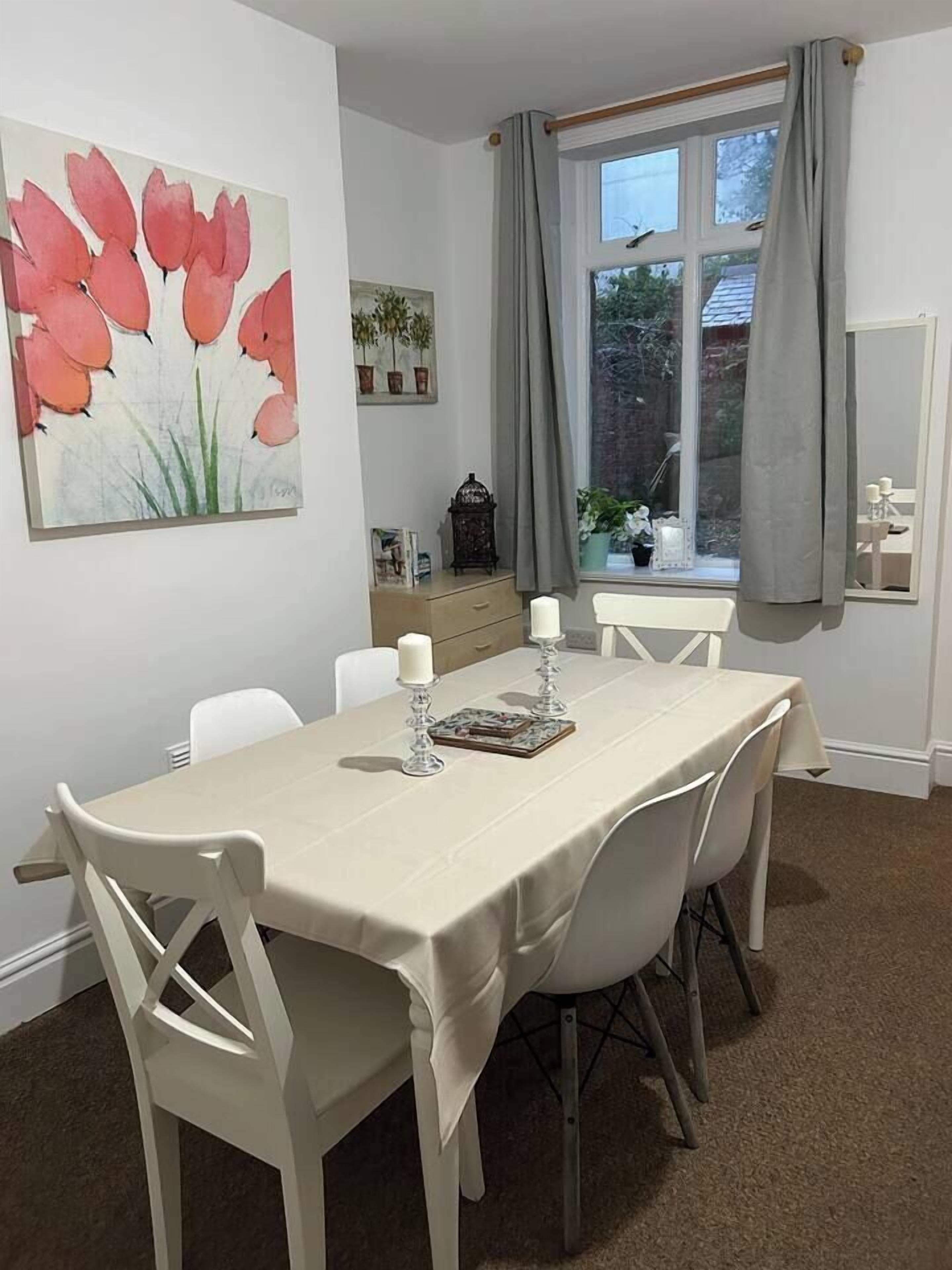 A dining room with a table set for four, a painting on the wall, and a window with curtains.