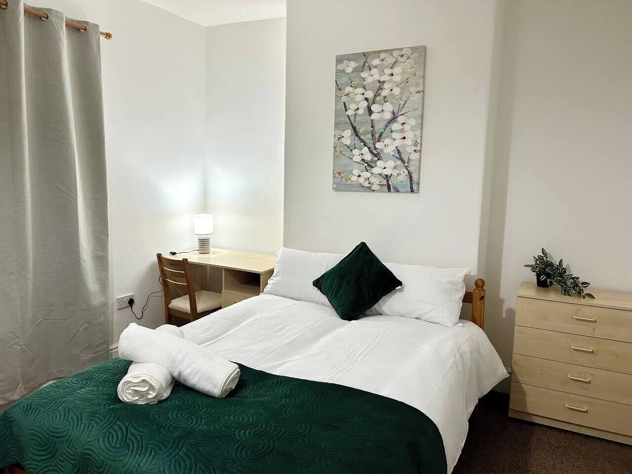 A neatly made bed with white bedding and a green throw, a desk with a chair, a dresser, and a painting on the wall.
