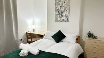 A neatly made bed with white bedding and a green throw, a desk with a chair, a dresser, and a painting on the wall.