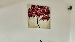 En snyggt bäddad säng med en röd filt och kuddar, ett sängbord med en lampa och en växt, och en tavla med ett träd som hänger på väggen.
