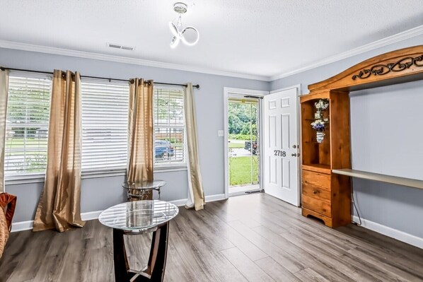 A room with a wooden floor, a glass table, curtains, a door with a number, and a cabinet.