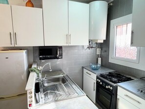 A modern kitchen with white cabinets, a built-in microwave, a sink with a dish rack, a stove, and a window with a view of trees. - House with 2 bedrooms and a shared pool (Empuriabrava)