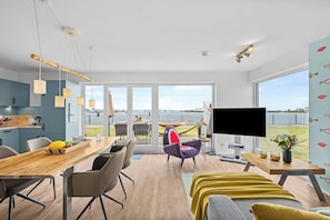 A modern living room with a large window, a flat-screen TV, and a dining area with a wooden table and chairs. - Fischerman's Friend - First row on the waterfront, infrared sauna, 3 TVs, close to the beach (Olpenitz)