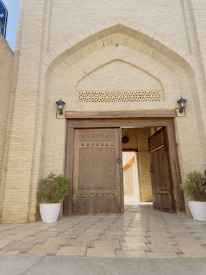 A large wooden door with intricate carvings, flanked by two potted plants, leading into a building with a brick facade and an arched entrance.
