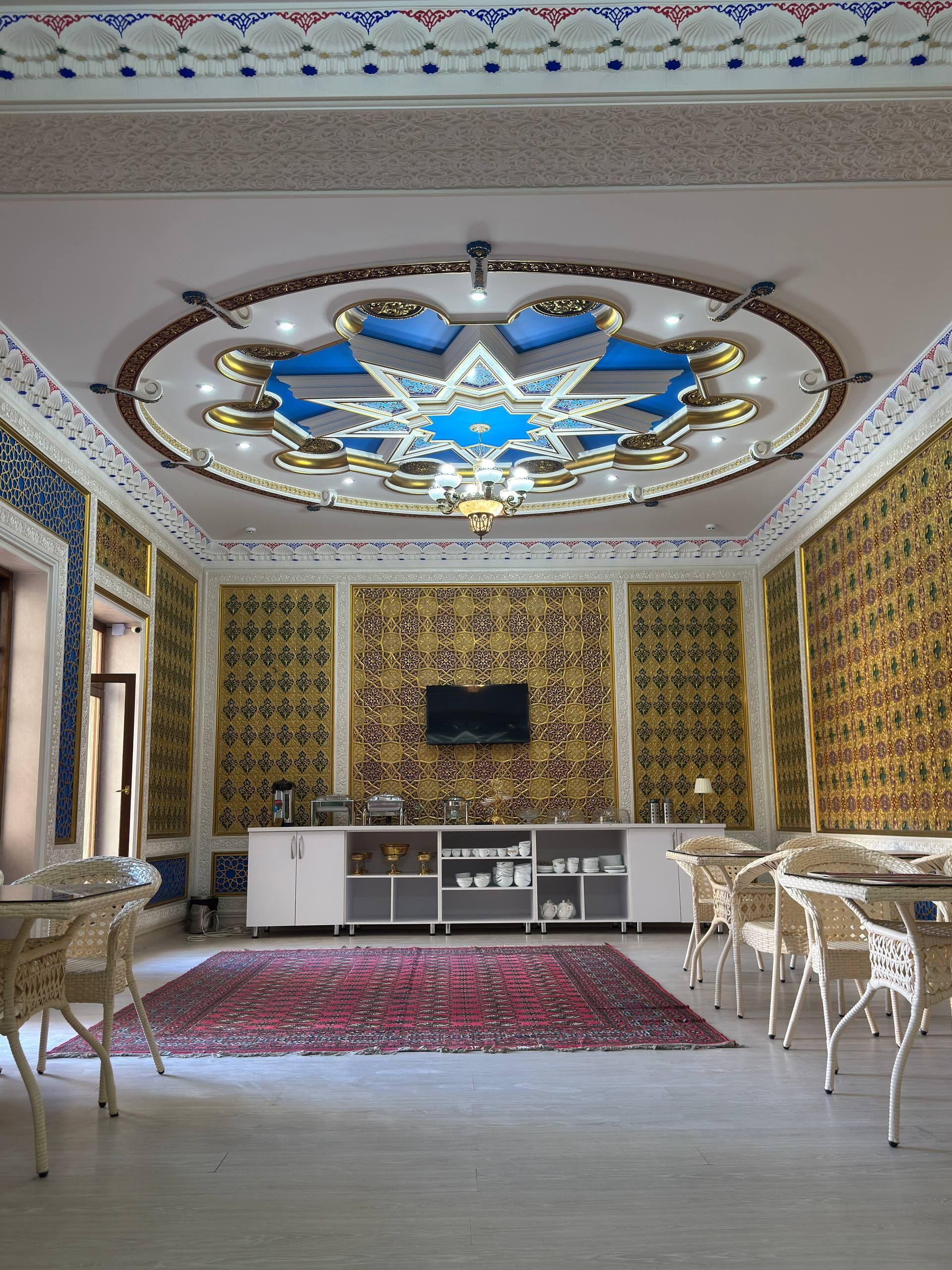 Una habitación con techo en forma de estrella, un televisor de pantalla plana montado en la pared, una consola blanca con varios objetos, sillas de mimbre y una alfombra estampada.