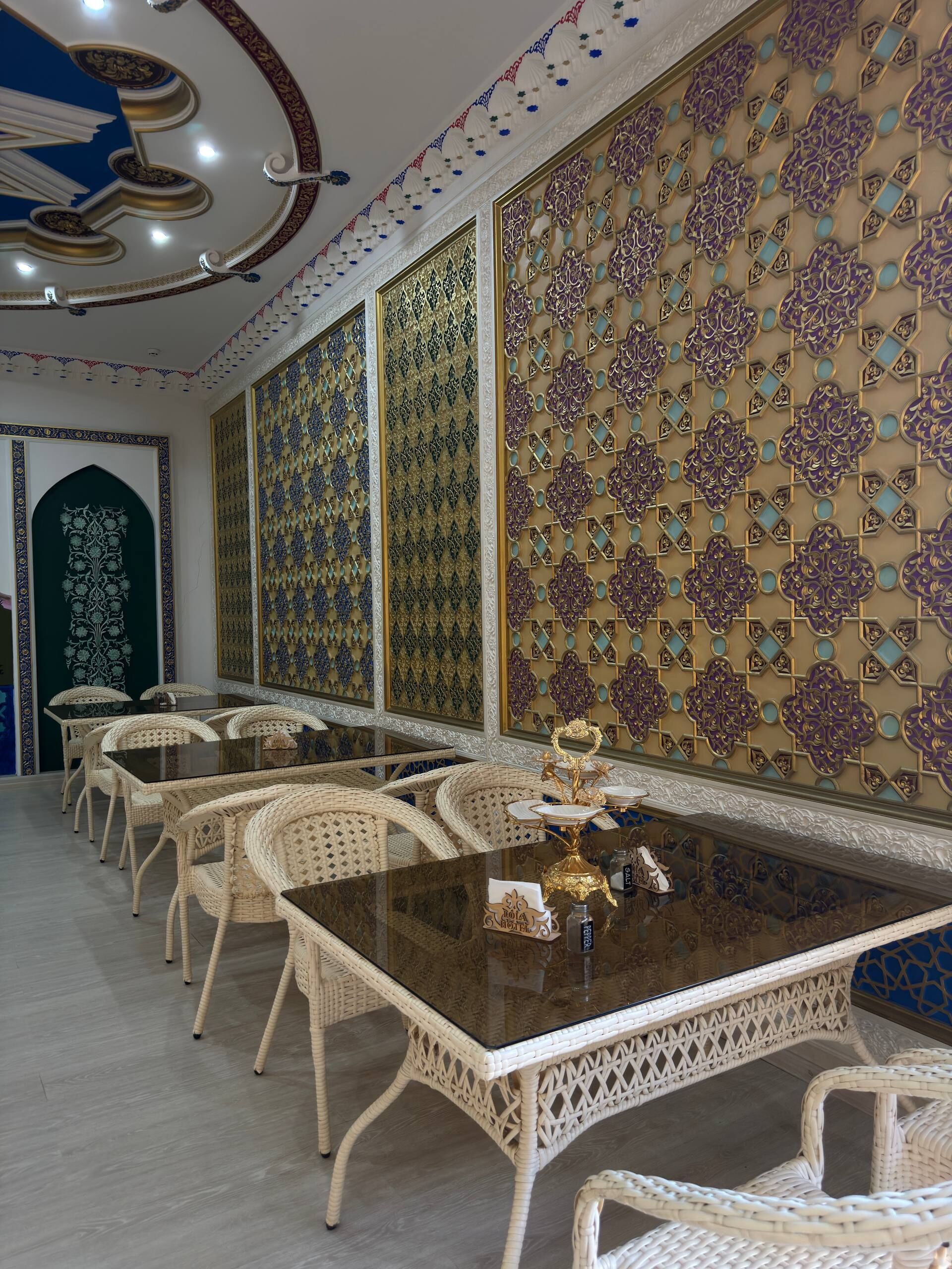 Un lungo tavolo da pranzo con sedie in vimini bianco, circondato da pannelli murali decorati con motivi elaborati.