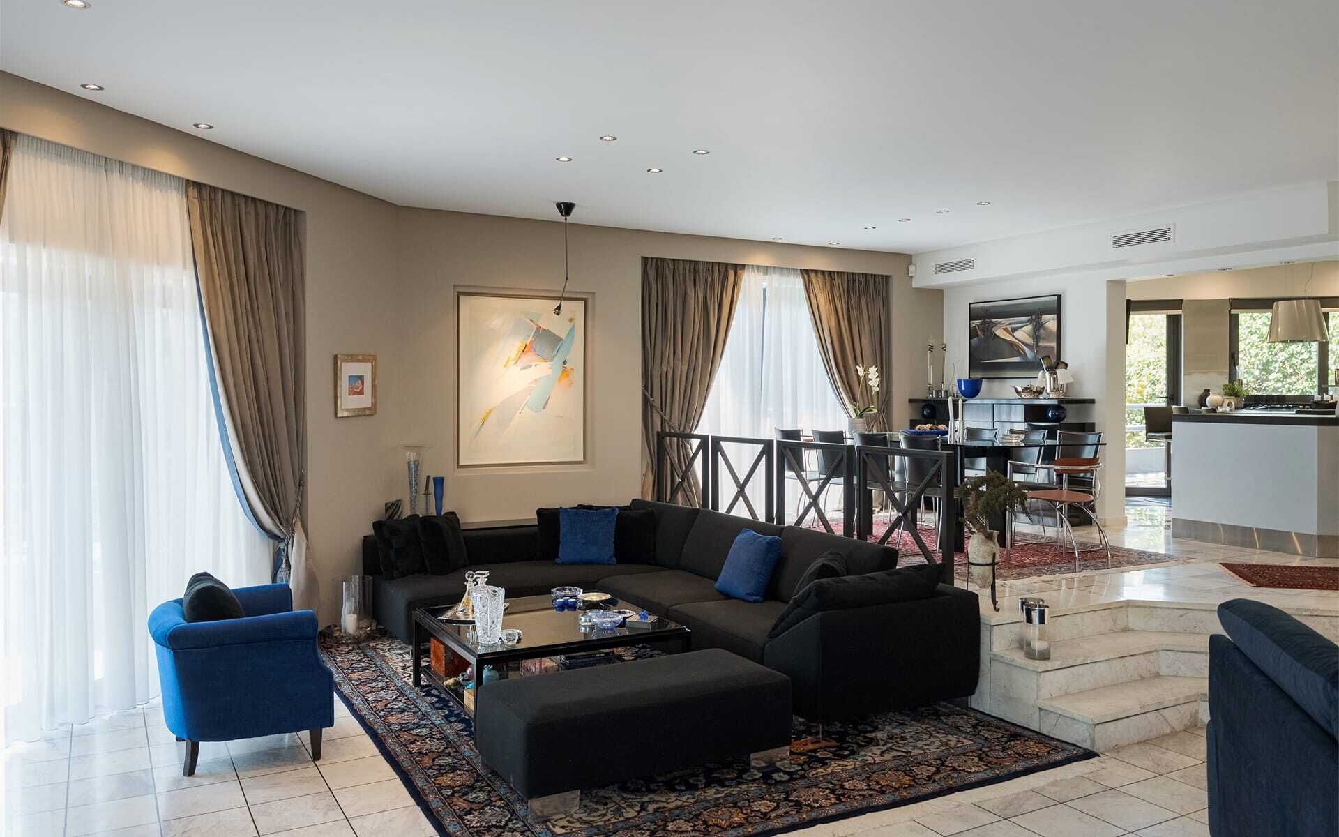 A modern living room with a black sofa, blue armchairs, and a coffee table. There is a dining area with a black table and chairs, and a kitchen visible in the background.