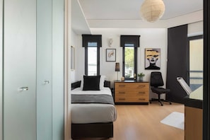 A modern bedroom with a bed, a desk, and a chair. There is a window with a view of trees.