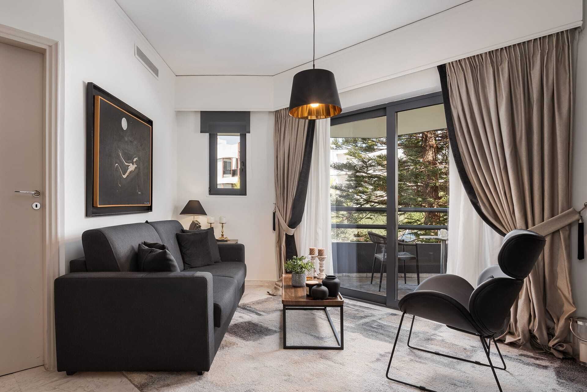 Un salon moderne avec un canapé, une table basse et un fauteuil. Il y a une grande fenêtre avec des rideaux, un luminaire suspendu et un tableau encadré au mur.