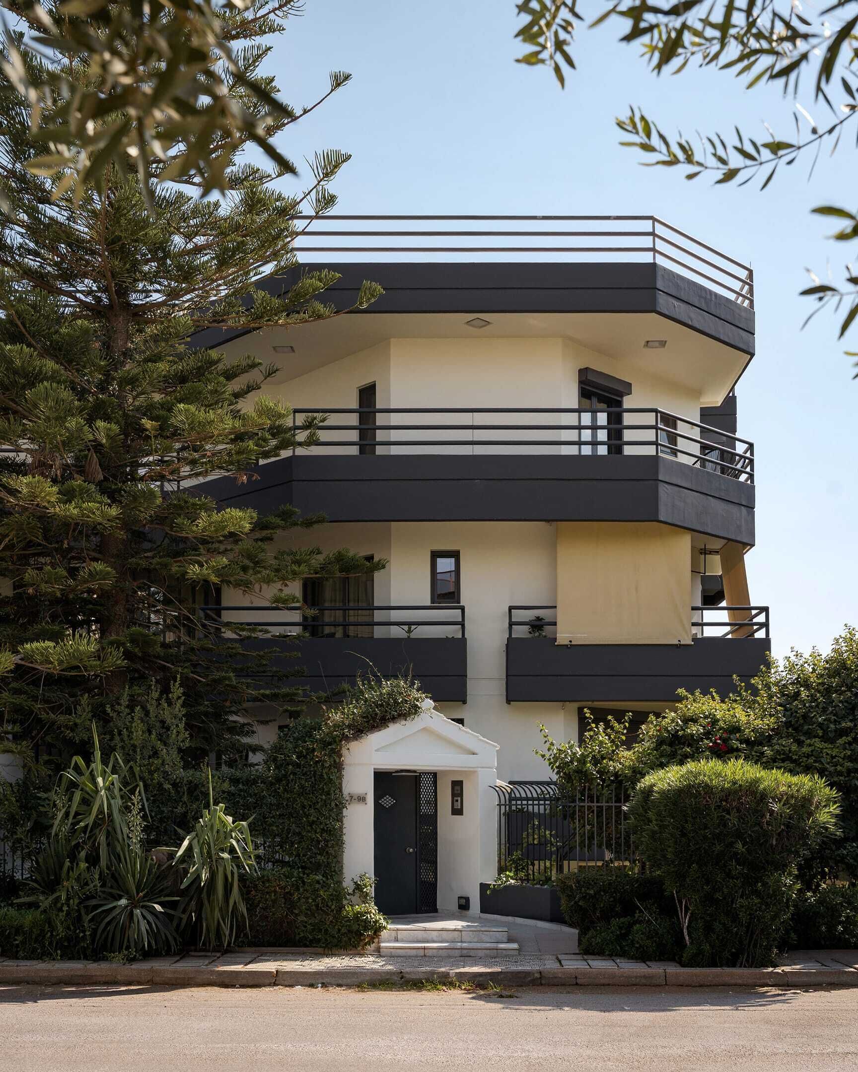 A multi-story residential building with balconies, a small entrance, and surrounding greenery.