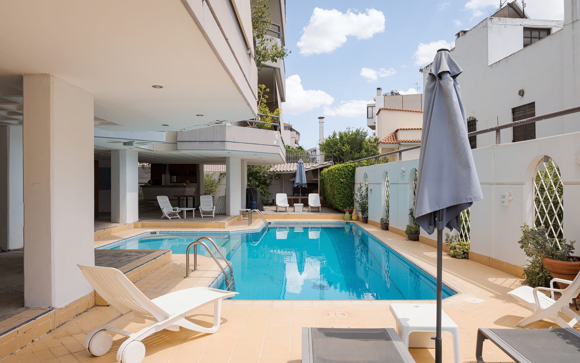 A swimming pool with sun loungers and a covered seating area, surrounded by a white building with a balcony.