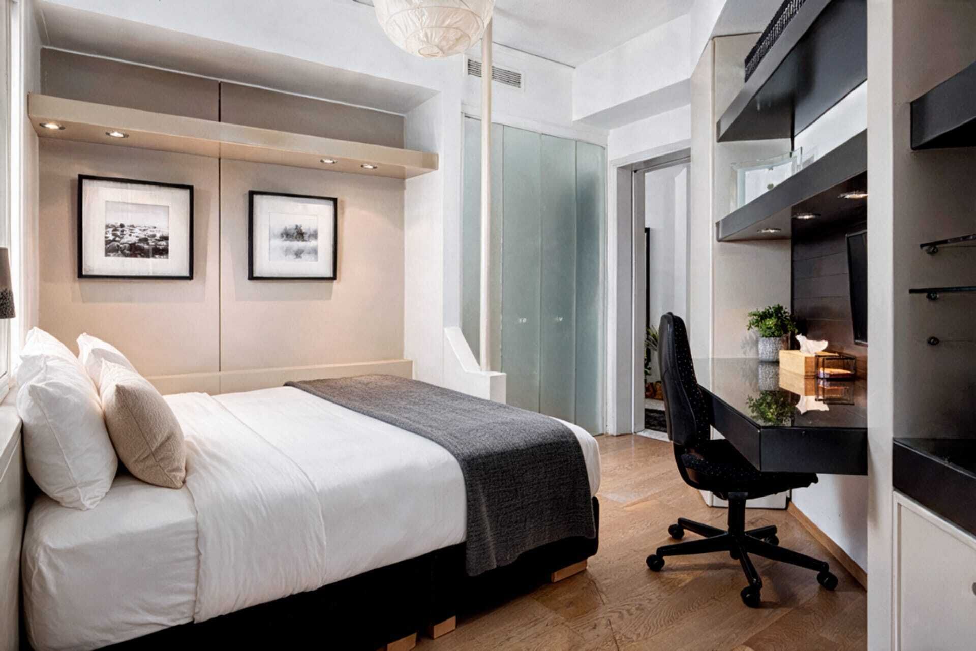 A modern bedroom with a bed, a desk, and a chair. There are two framed pictures on the wall.