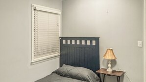 A single bed with a headboard, a bedside table with a lamp, a window with blinds, and a light switch on the wall. - Charming Single-Family Home (Greenville)
