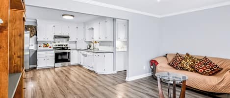 A modern kitchen with white cabinets and a central island, a wooden floor, and a beige sofa with patterned cushions.
