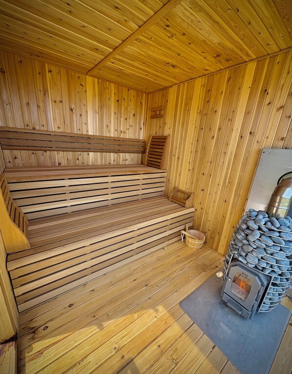 A wooden sauna with benches, a stone-filled container, and a wood-burning stove.