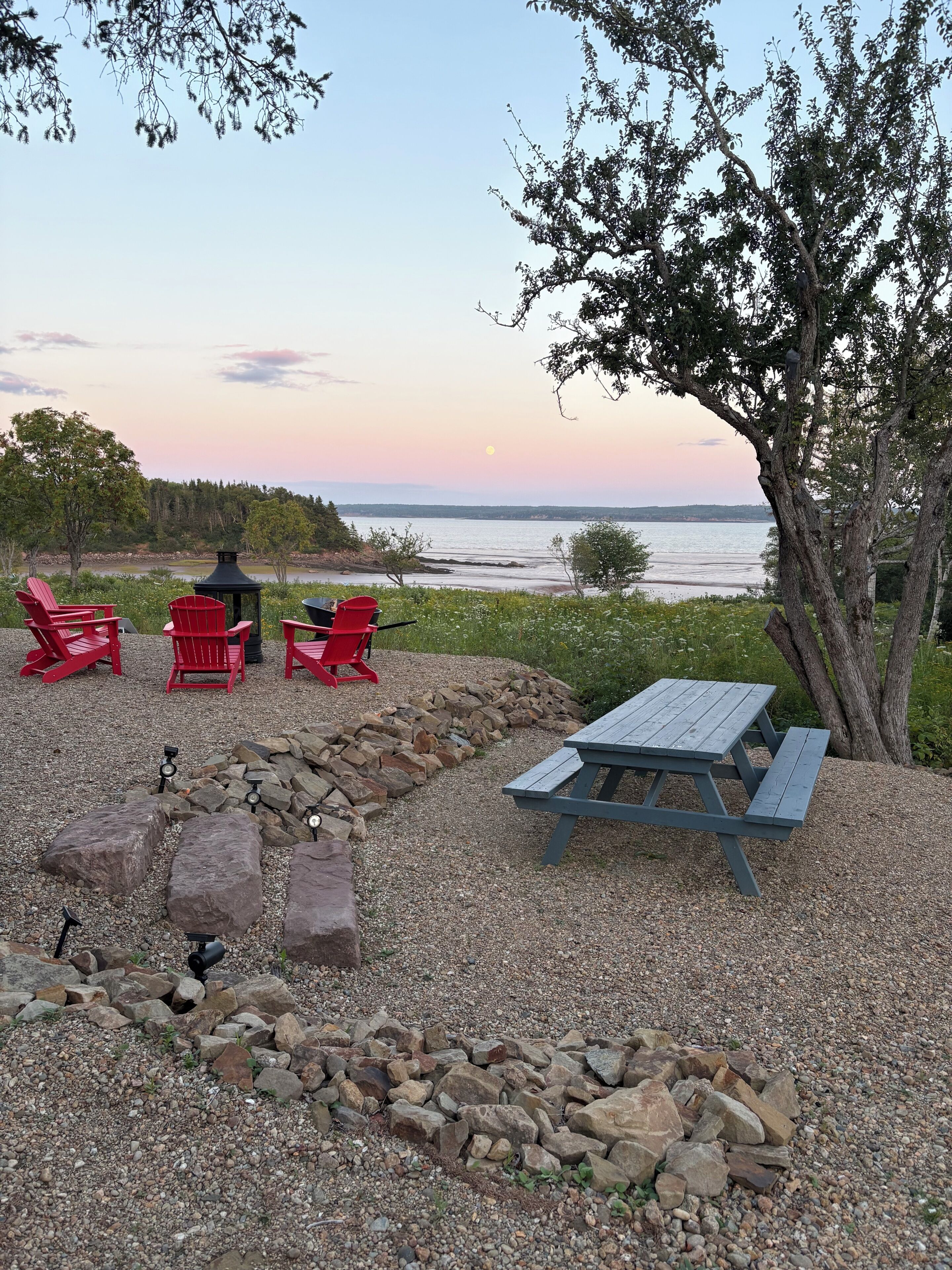 A gravel patio with a picnic table and red chairs, overlooking a body of water.