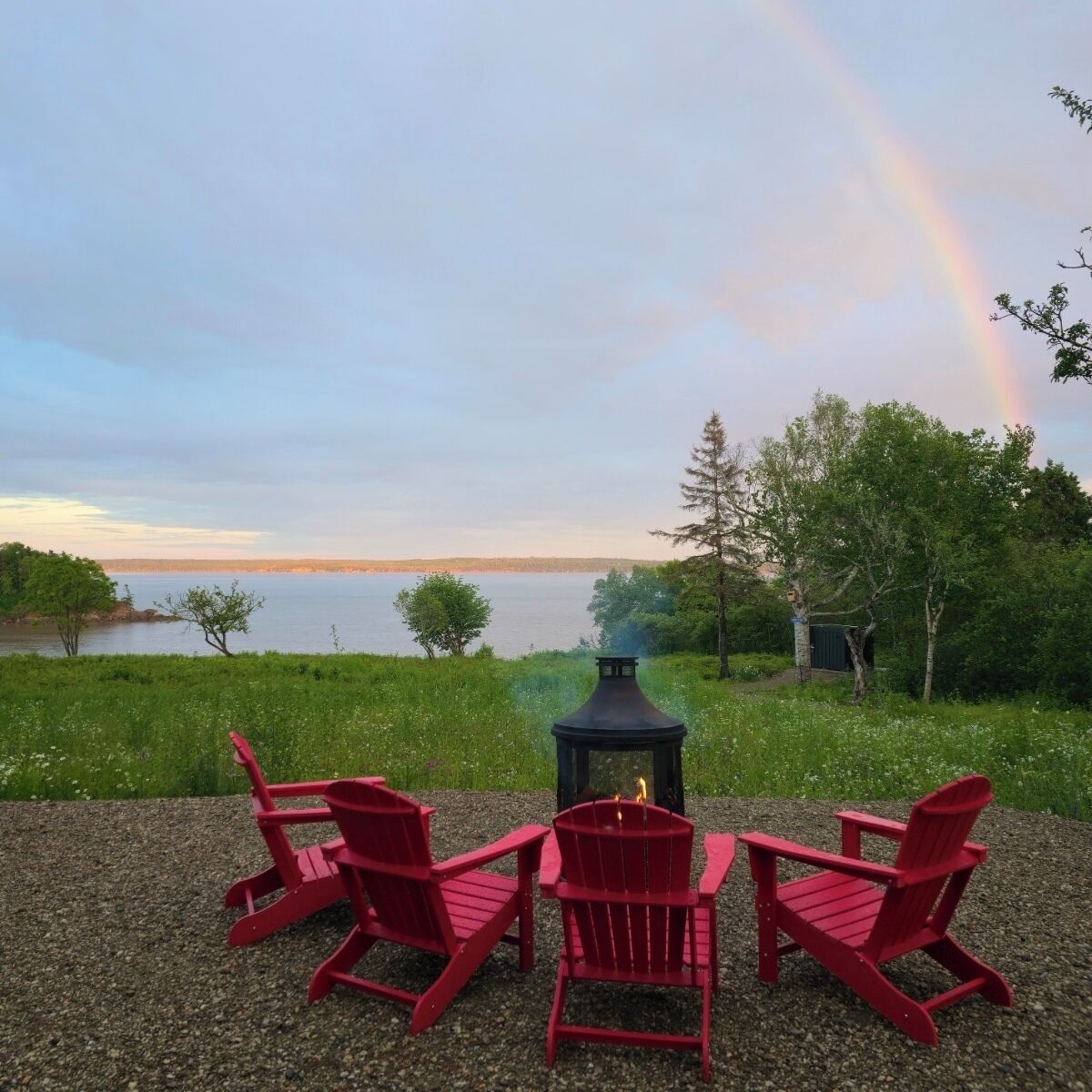 Eine Seenlandschaft mit Feuerstelle und roten Adirondack-Stühlen.