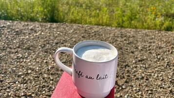 Une tasse blanche avec du texte sur une surface rouge, placée à l’extérieur, avec un paysage naturel en arrière-plan.