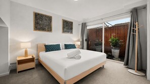 Ett hotellrum med en säng, ett nattduksbord, en lampa, konstverk och utsikt genom en skjutdörr i glas.
