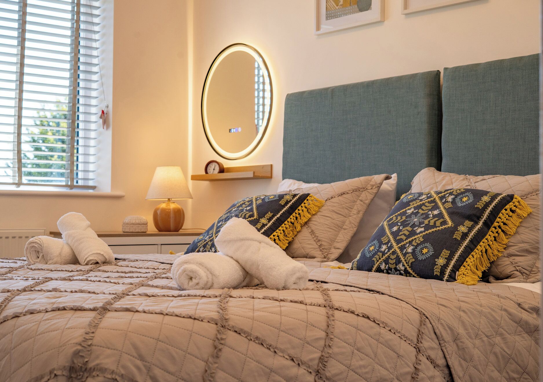 A neatly made bed with rolled towels, a bedside lamp, and a wall-mounted mirror.