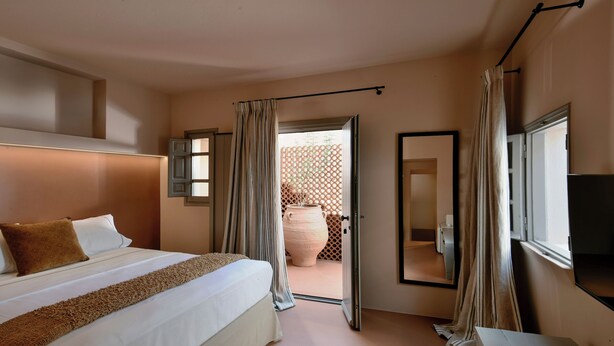 A hotel room with a large bed, a flat-screen TV, a window with curtains, and a view of the outside. - Terra Verde Oia Suites - Cotto Standard Room (Oia)