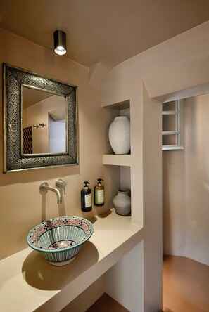 A bathroom with a decorative mirror, a bowl sink, and shelves with toiletries. - Terra Verde Oia Suites - Cotto Standard Room (Oia)