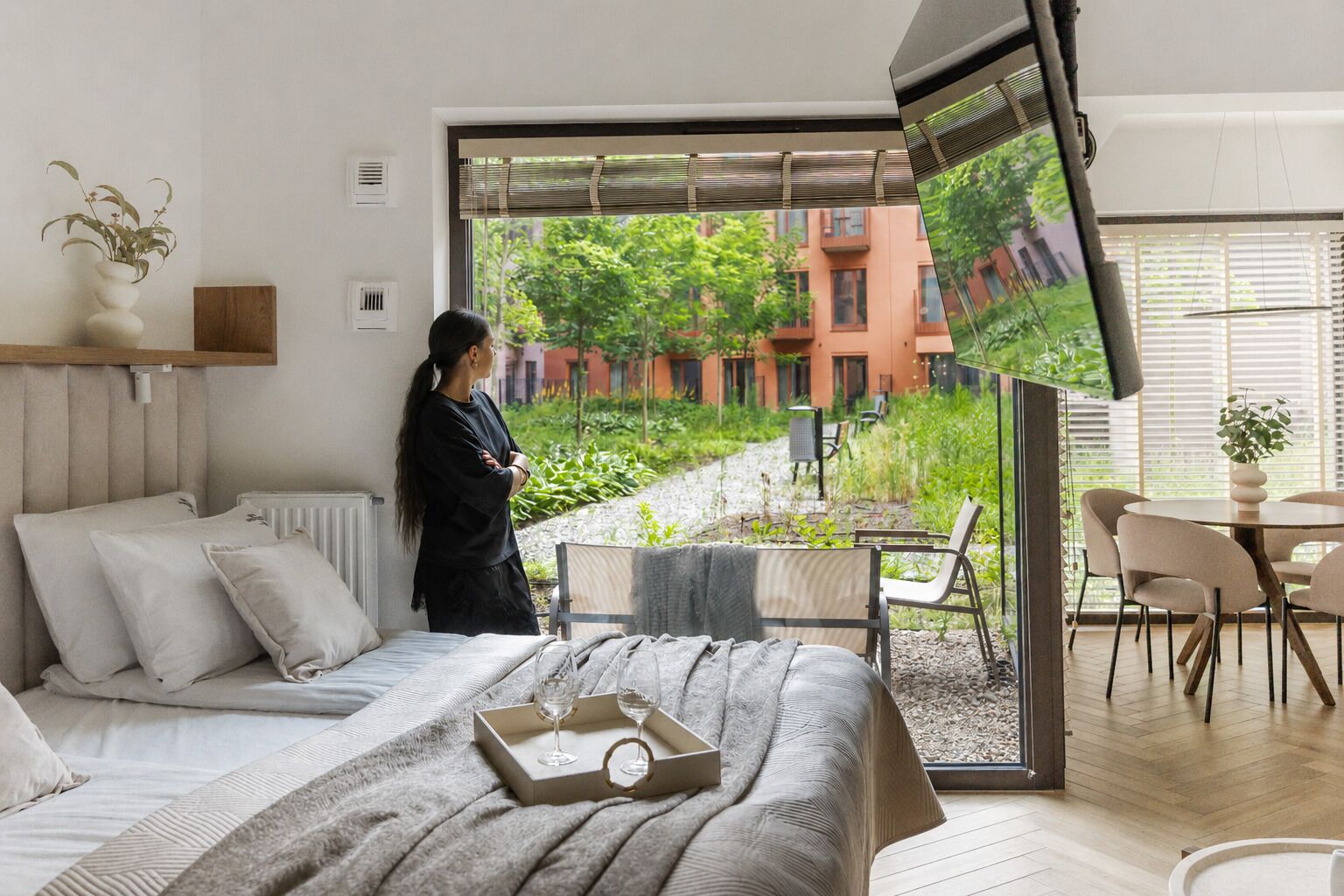 A bedroom with a bed, bedside table, and a large window with a view of greenery and buildings.