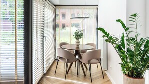 Ein modernes Interieur mit einem runden Tisch und Stühlen, großen Fenstern mit Jalousien und einer Zimmerpflanze.