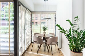 Ein modernes Interieur mit einem runden Tisch und Stühlen, großen Fenstern mit Jalousien und einer Zimmerpflanze.