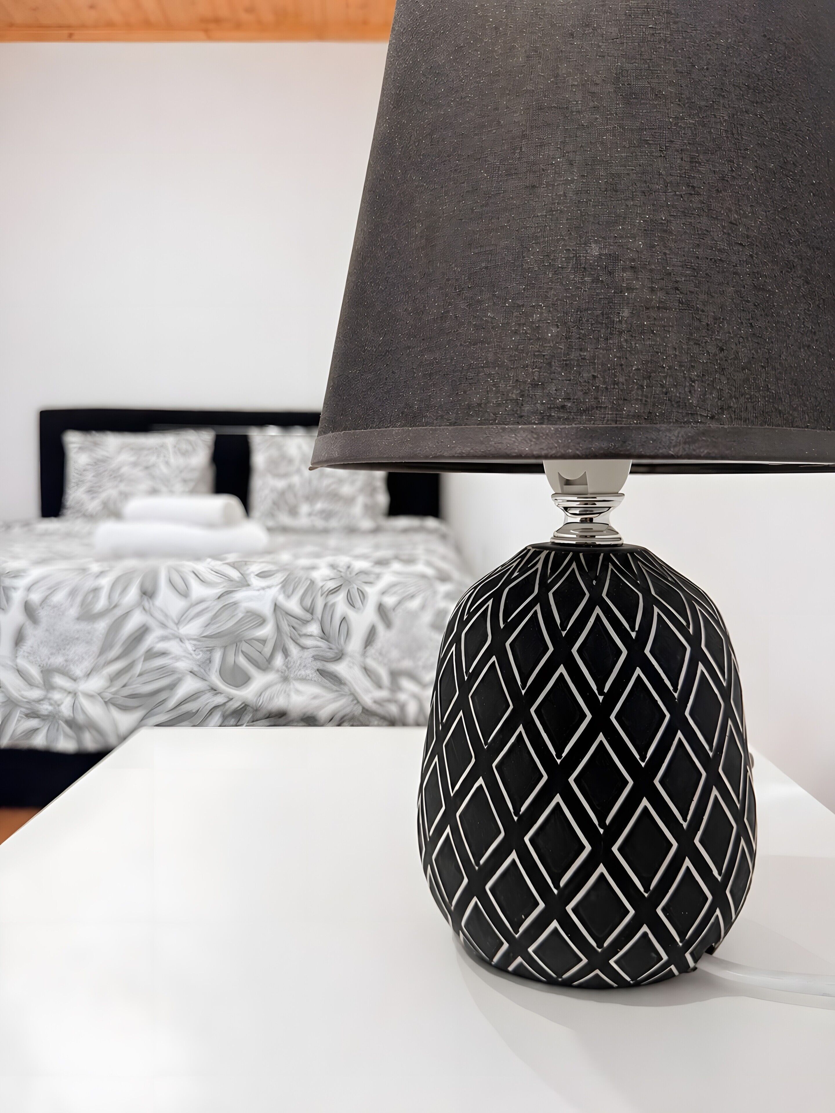 A lamp with a geometric pattern on a white surface, a bed with a patterned bedspread, and a plain wall in the background.