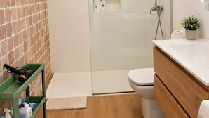 Et moderne badeværelse med en glasbrusekabine, toilet, håndvask med spejl og en grøn hylde med toiletartikler.