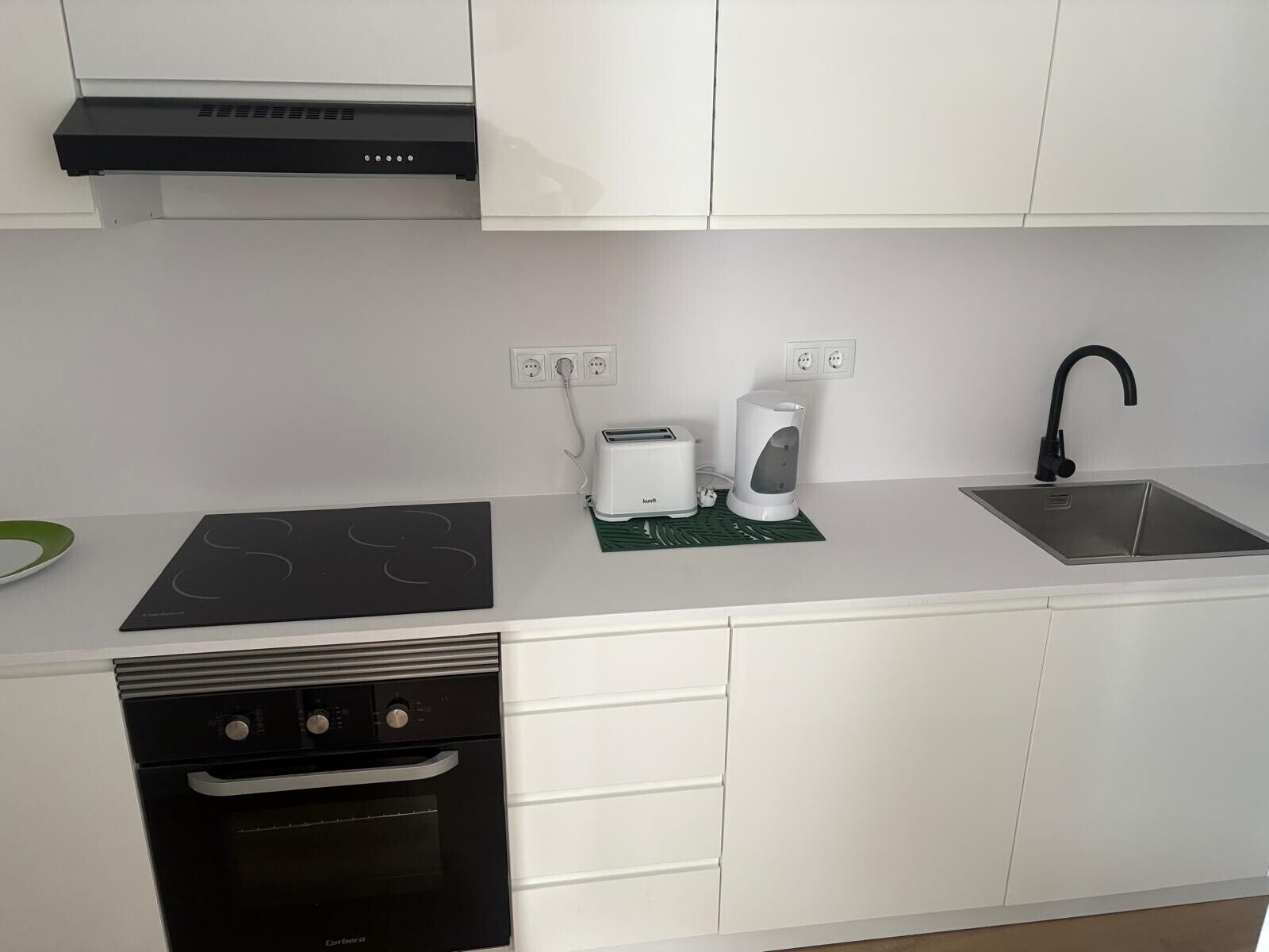 A modern kitchen with white cabinets, a black dishwasher, a built-in oven, a stainless steel sink, and a toaster.