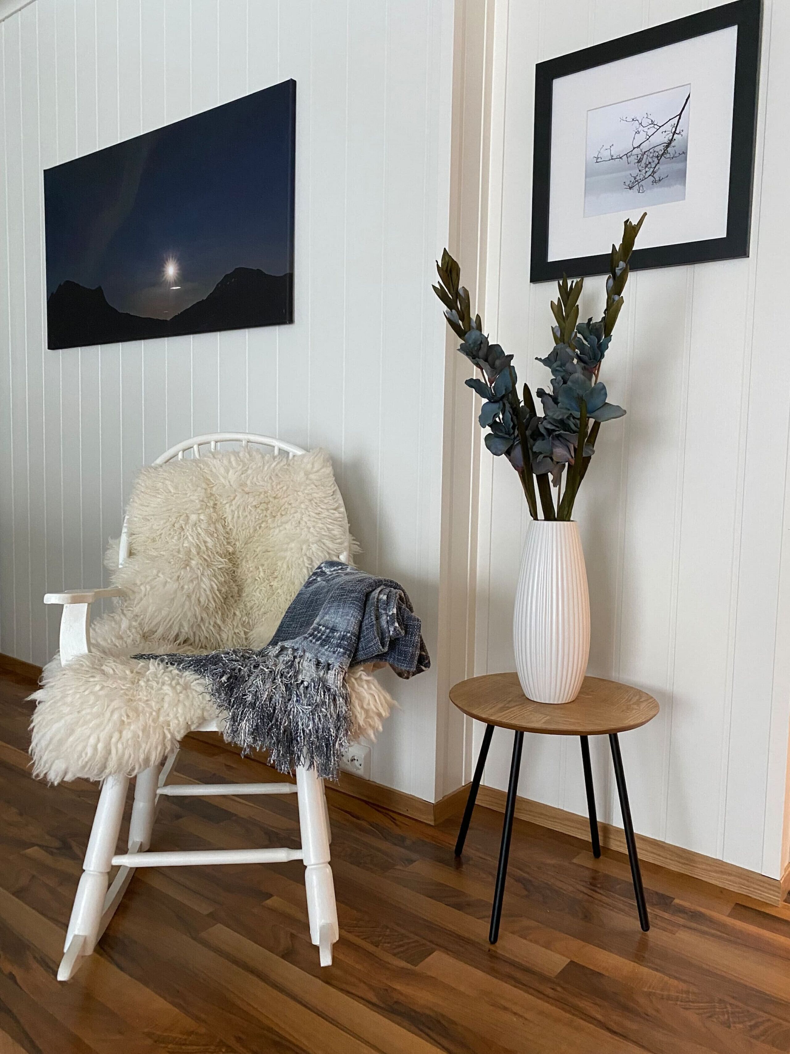 A room with a white chair with a fluffy cushion, a small round table with a vase of blue flowers, and two framed pictures on the wall.