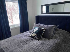 Una cama con cubrecolcha gris acolchado, dos almohadas con motivos florales, un cabecero oscuro y una ventana con cortinas azules.