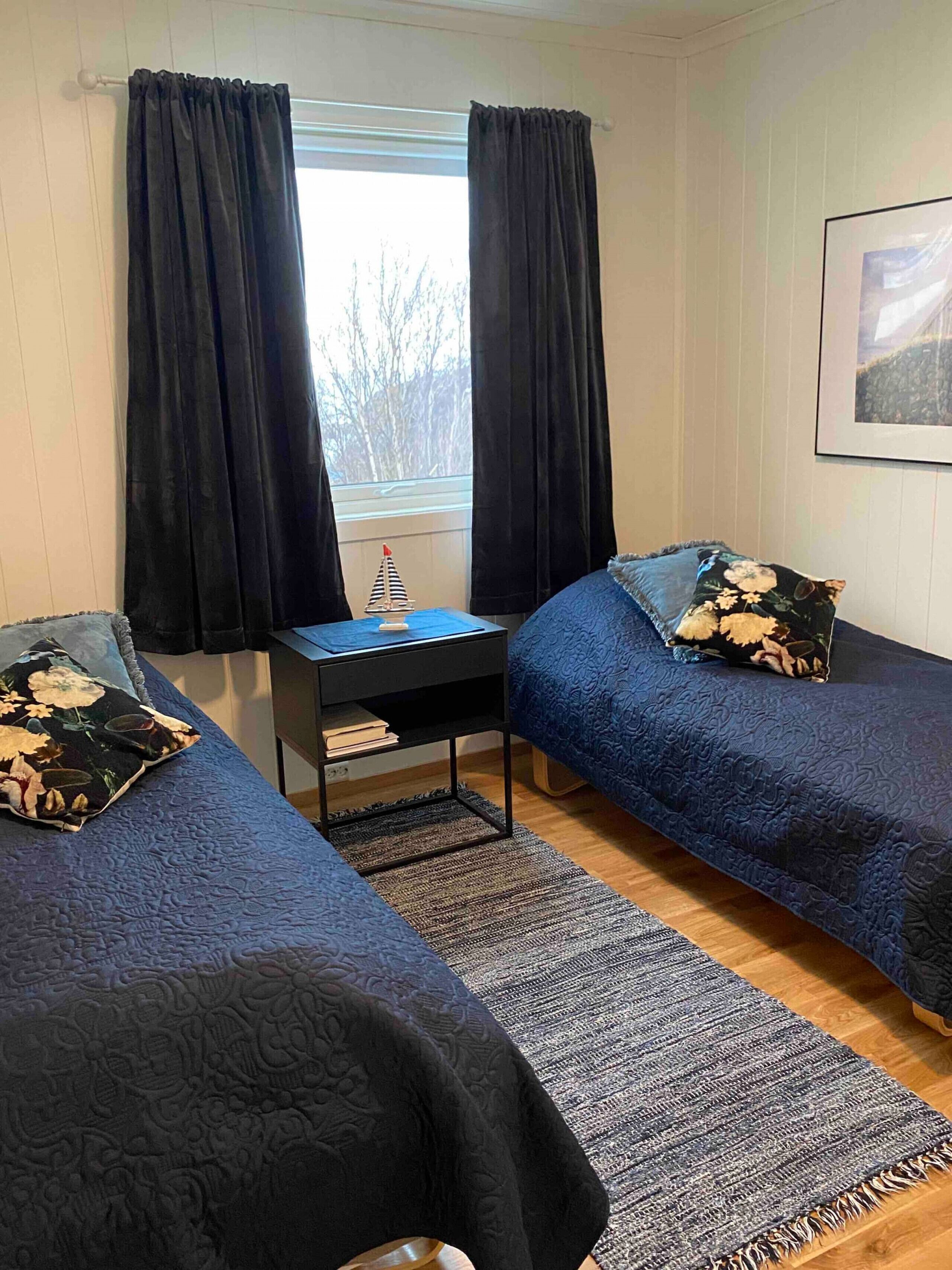 A room with two beds, a small table, a rug, and a window with curtains.