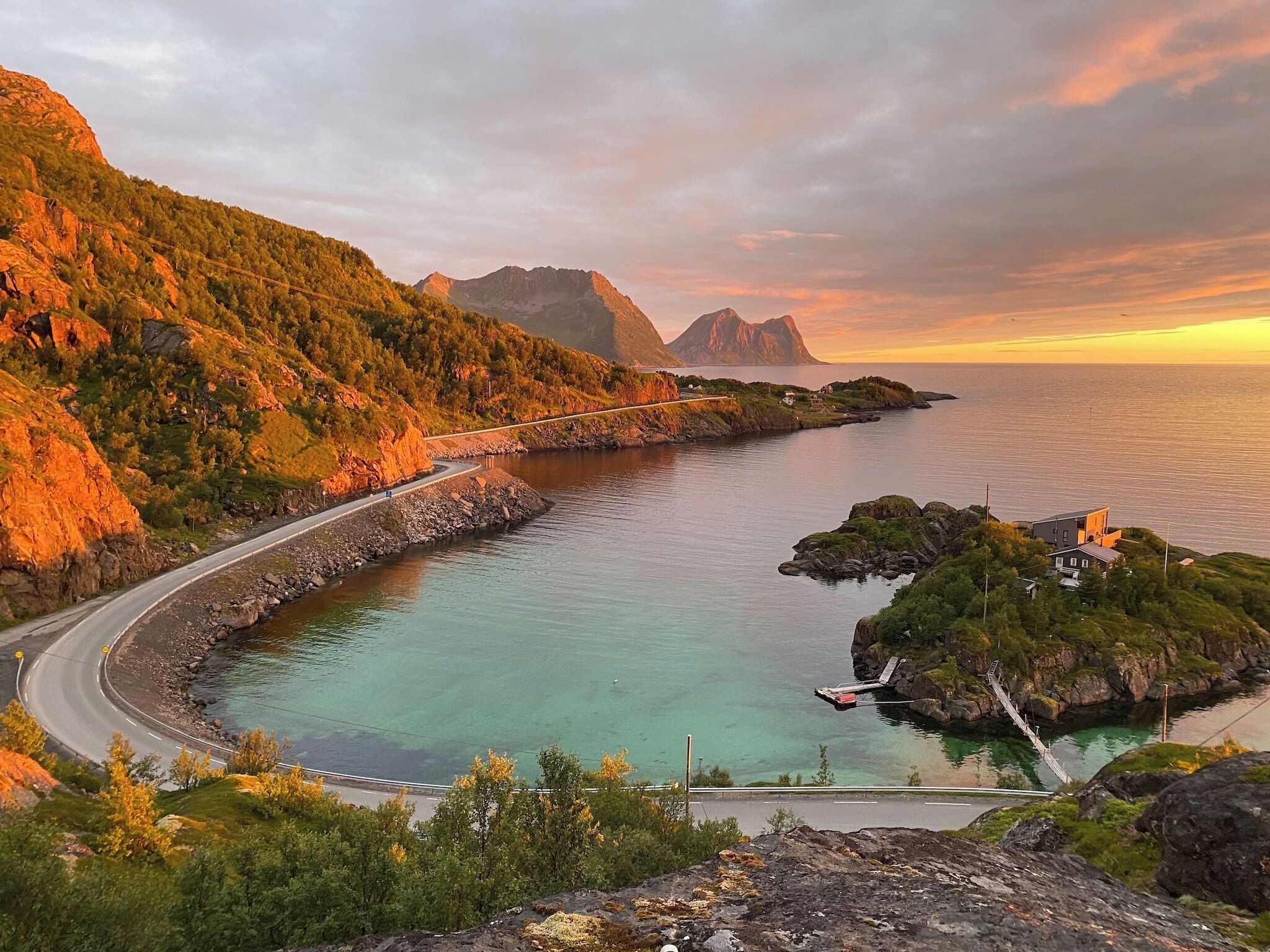 A coastal road curving along a bay with clear water, surrounded by green hills and a mountain in the distance.