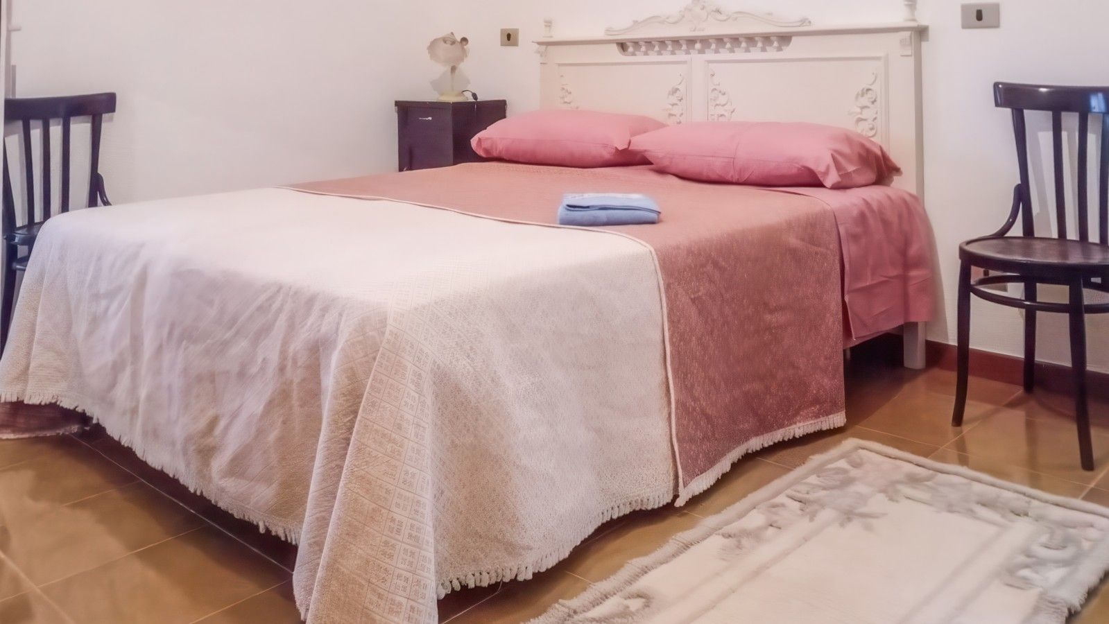Una cama con una colcha blanca y rosa, una mesita de noche con una lámpara y una silla.