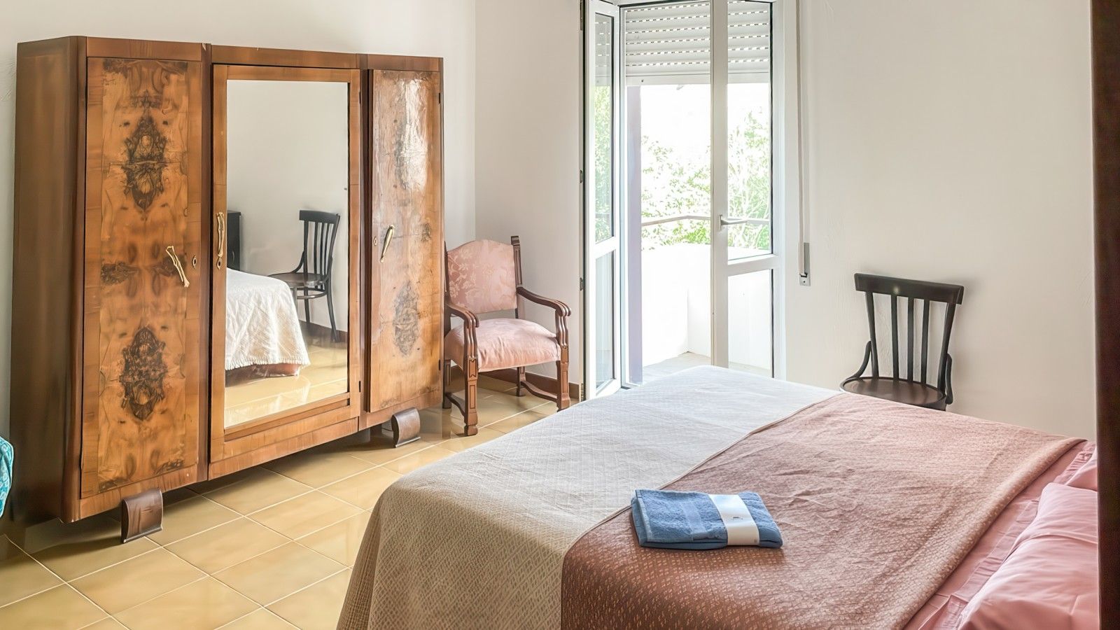 Una camera da letto con un armadio capiente, un letto con copripiumino rosa e bianco, una sedia e una porta scorrevole in vetro che dà su un balcone.