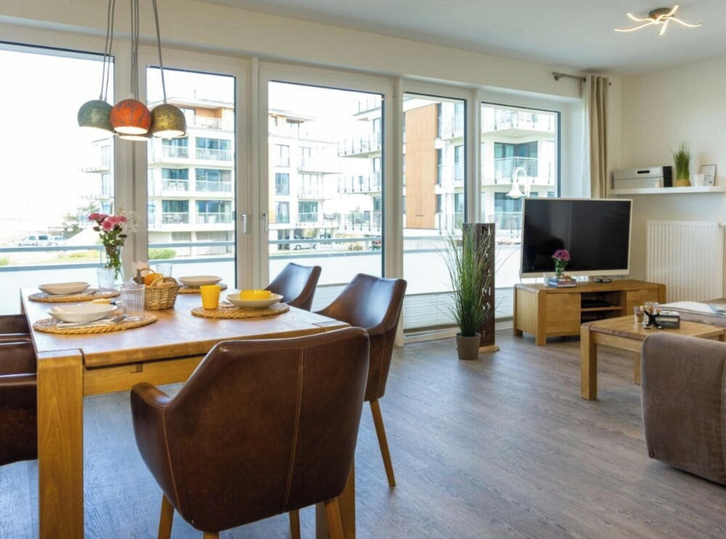 A modern living room with a dining area, large windows, a flat-screen TV, and a potted plant.