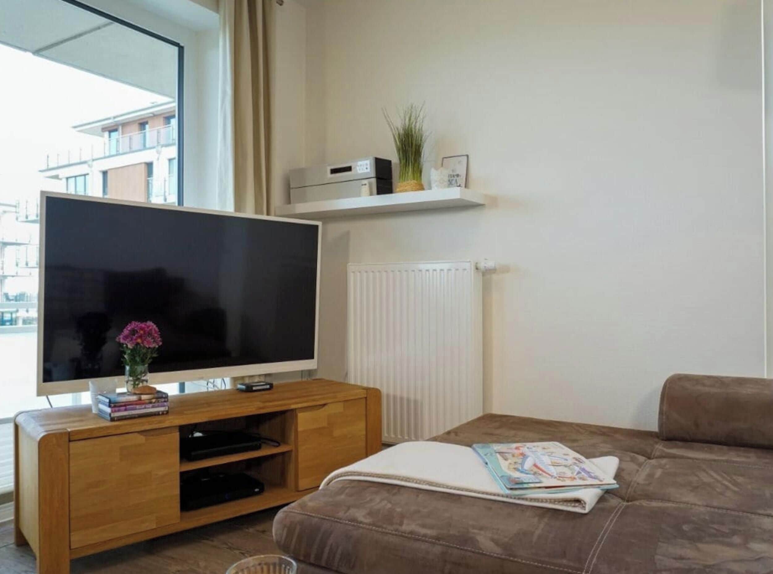 A modern living room with a large flat-screen TV on a wooden stand, a brown sofa, a small table with a magazine, and a radiator.