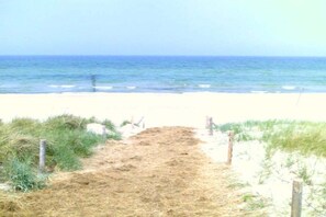 Ein sandiger Küstenpfad führt zum Meer, begleitet von spärlicher Vegetation an beiden Seiten.